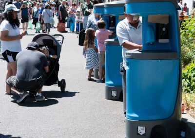 A row of blue multi-user portable hand washing stations being used by families at a crowded outdoor festival.