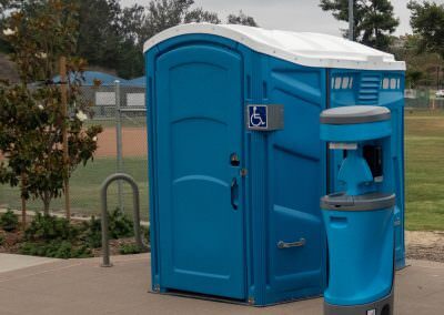 A blue wheelchair-accessible portable restroom paired with a compact external hand wash station at a community sports field.
