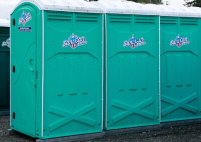 Three teal-colored standard portable restroom units lined up on a gravel surface for outdoor event sanitation.